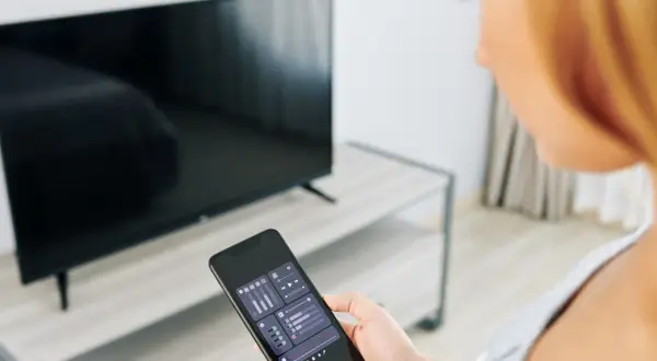 Kobieta trzyma smartfon z aplikacją do sterowania TV, obok stoi włączony telewizor.
